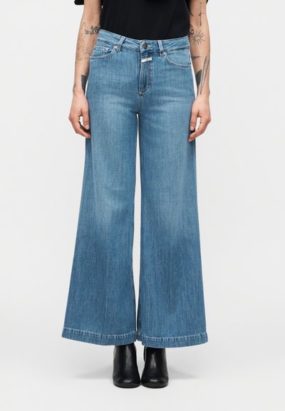 Personne portant un jean taille haute à jambes larges de couleur bleue, associé à des bottines noires et un haut noir, debout devant un fond uni.