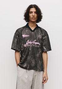 Sort sportsjersey med et marmoreret mønster, pink tekst "Asha Square Sports Club" og en kontrasterende sort V-halskrave. Korte ærmer.