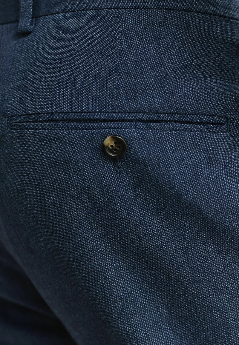 Primo piano di pantaloni in tessuto blu scuro che mostrano una tasca posteriore a filetto chiusa con un singolo bottone scuro.