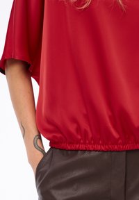 Arm met slangtattoo, hand in de zak, draagt een losse rode blouse met elastische zoom en donkerbruine leren broek.
