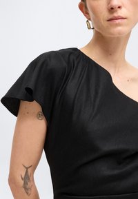 Γυναίκα που φοράει μαύρο τοπ με έναν ώμο και βολάν στο μανίκι, χρυσό κρίκο σκουλαρίκι και ορατά τατουάζ στο χέρι με ψάρι και ένα θλιμμένο emoji.