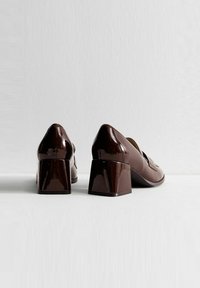 Tacones de charol marrón con un tacón cuadrado y punta redondeada. Superficie lisa con un mínimo de detalles y sin herrajes visibles.