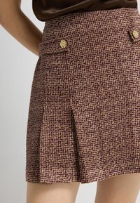 Jupe courte plissée en tissu texturé avec un motif bordeaux et beige. Présente des boutons dorés à la taille.