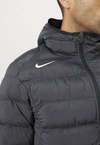 Sort puffersjakke med hætte har syede sektioner og en frontlynlås. Hvidt Nike-logo på venstre skulder. Glat, skinnende tekstur.