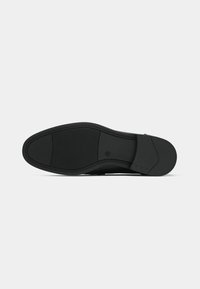 Semelle de chaussure noire avec une texture lisse, un bord légèrement surélevé et un motif antidérapant pour la traction. Présente un marquage discret près du talon.
