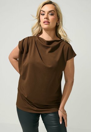 Blondynka w brązowym, drapowanym topie z krótkim rękawem, połączonym z czarnymi skórzanymi spodniami, stoi z jedną zgiętą ręką na neutralnym tle.