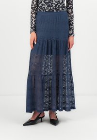 Falda maxi de encaje azul oscuro, con un diseño en capas y bordes en forma de ondas. Combinada con unos zapatos negros de tacón abierto y puntera afilada.