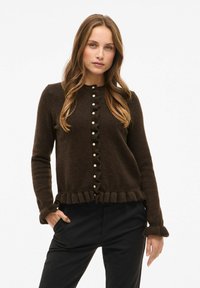 Brūns adīts kardigans ar viļņotu priekšpusi, pērļu pogām un frillētiem piedurkņu galiem. Tam ir mīksta tekstūra un pieguļošs dizains.