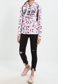 Felpa grigia con motivo floreale, tasca a marsupio, jeans neri strappati e sneakers rosa chiaro. Cappuccio con lacci e testo del logo.