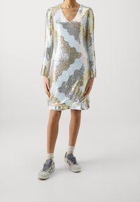 Hedvábné šaty se V výstřihem a dlouhými rukávy, zdobené vlnitým vzorem v zlaté, stříbrné a modré barvě. Kombinované s šedými teniskami a krémovými ponožkami.