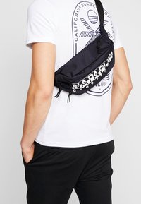 Uomo che indossa una t-shirt bianca con grafica e pantaloni neri, con una borsa a tracolla nera Napapijri sulla schiena.