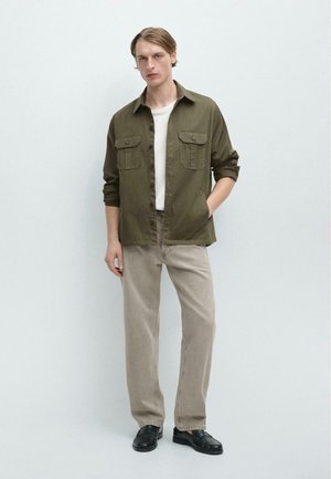 Jonge man met een olijfgroen overhemd over een wit T-shirt, beige broek en zwarte loafers, staand met één hand in de zak tegen een witte muur.