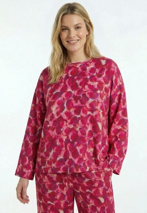 Frau trägt passendes langärmeliges Oberteil und Hose mit pink-rot abstraktem Blumenmuster, lächelt vor schlichtem Hintergrund.