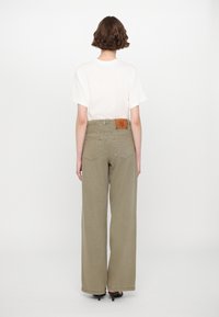Weite khakifarbene Hose mit hoher Taille, einer aufgesetzten Gesäßtasche und einem braunen Lederetikett auf der Rückseite. Kombiniert mit einem schlichten weißen T-Shirt.