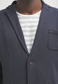 Hombre con blazer azul oscuro sobre una camiseta de rayas blancas y grises con cuello redondo, abotonada al frente.