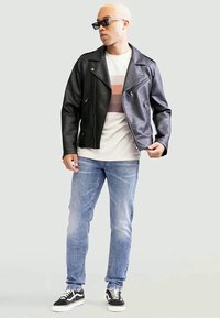 Casaco de couro preto estilo biker com ferragens prateadas, por cima de uma t-shirt branca às riscas. Par de jeans azul claro e ténis pretos.