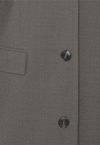 Grijze blazer met een gladde textuur, voorzien van een klepzak en een ronde zwarte knop met een decoratief ontwerp in het midden.