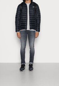Black puffer jacket with horizontal stitching, grey t-shirt, grey jeans, and black sneakers. Backpack straps visible on shoulders.