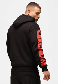 Felpa nera con un design stampato rosso sulla manica sinistra, caratterizzata da una grande scritta "GREETING". Il materiale sembra morbido; include un cappuccio regolabile con cordino.