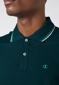 Polo shirt verde scuro in tessuto testurizzato, con tre bottoni e colletto a contrasto con righe verdi e bianche. Include un logo ricamato.