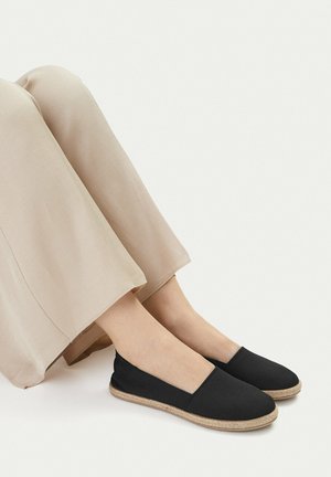 Nogi w beżowych spodniach i czarnych wsuwanych espadrylach z plecionymi jutowymi podeszwami, przedstawione na jednolitym tle.