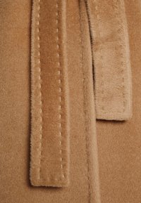 Bruine fluweelstof met twee langwerpige, gestikte strikken. De textuur is zacht, met gouden draad voor detailing langs de randen.