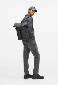 Μαύρος σάκος πλάτης με σχεδίαση ρολού και ρυθμιζόμενους ιμάντες. Το μοντέλο φοράει ένα γκρι τζάκετ από τζιν και αντίστοιχα γκρι τζιν παντελόνι, με μαύρα παπούτσια.