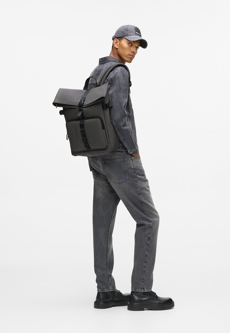 Μαύρος σάκος πλάτης με σχεδίαση ρολού και ρυθμιζόμενους ιμάντες. Το μοντέλο φοράει ένα γκρι τζάκετ από τζιν και αντίστοιχα γκρι τζιν παντελόνι, με μαύρα παπούτσια.