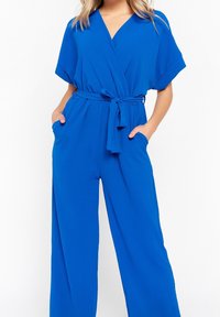Kombinezon w królewskim niebieskim kolorze, z zapięciem w stylu kopertowym, krótkimi rękawami, paskiem do związania w talii i bocznymi kieszeniami. Wykonany z gładkiego materiału; szerokie nogawki zwiększają komfort.