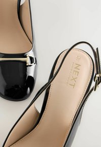Musta lakattu nahka slingback-korkokengät, joissa on koristeellinen kultabuketti, korostavat neutraalia beigen sisäpohjaa, johon on painettu brändin nimi.