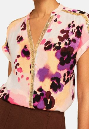 Vrouw draagt een blouse met korte mouwen met een roze, paarse en zwarte abstracte bloemenprint en gouden bies langs de V-hals.