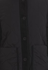 Veste matelassée noire avec un plastron en velours côtelé, cinq boutons et deux poches latérales. Texture lisse avec un motif ondulé subtil sur le tissu.