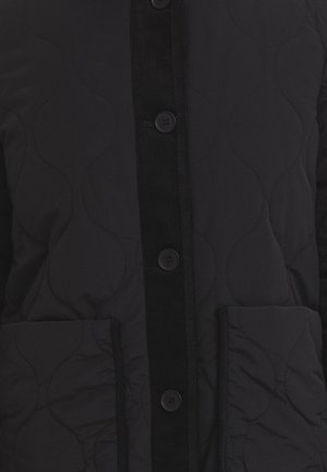 Schwarze gesteppte Jacke mit einem Cordura-Riegel, fünf Knöpfen und zwei Seitentaschen. Glatte Textur mit feinem Wellenmuster auf dem Stoff.
