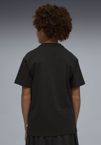 Άτομο με σγουρά μαλλιά, ορατό από πίσω, φορώντας μια απλή μαύρη κοντομάνικη μπλούζα και μαύρο υφασμάτινο παντελόνι, μπροστά σε ουδέτερο φόντο.