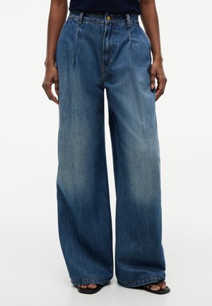 Person iført vidde, højtaljede blå denimjeans med falmede detaljer og sorte åbne sandal, hænder afslappet ned langs siden.