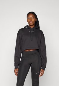 Svart cropped hettegenser med snor, med et logo i hvitt. Kombinert med høyhuskede svarte leggings med matchende logodetaljer.