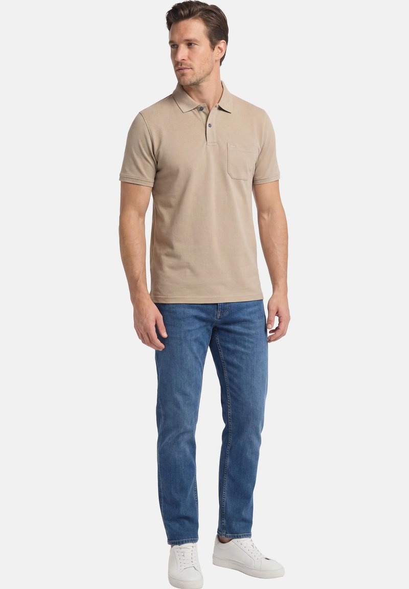 Mann steht und trägt ein beiges Poloshirt mit kurzen Ärmeln und Tasche, blaue Jeans und weiße Turnschuhe, schaut leicht zur Seite.