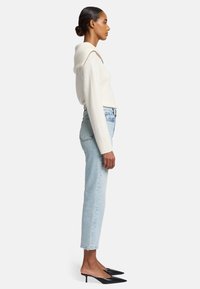 Maglione a coste crema con cappuccio, jeans azzurri a vita alta, e mule nere con tacco a punta, vista laterale su uno sfondo neutro.