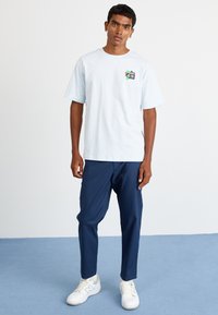 Lyseblå bomulds T-shirt med et grafisk kamera design på venstre bryst, parret med navy bukser og hvide sneakers. Enkel, afslappet stil.