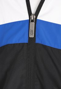 Schwarze und blaue Windjacke mit weißen Akzenten, ausgestattet mit einem zentralen Reißverschluss und einer glatten, glänzenden Textur. Der Reißverschluss-Zipper trägt das "Nike"-Logo.