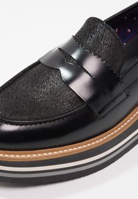 Mocassin en cuir noir avec dessus texturé, bride décorative, lisière marron et semelle en caoutchouc superposée noire et blanche.