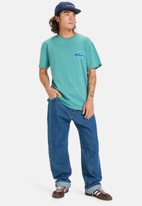 Camiseta de algodón verde azulado con un pequeño gráfico azul, combinada con jeans de mezclilla azul holgados y zapatillas de deporte marrones. El sombrero es navy con un logo blanco.