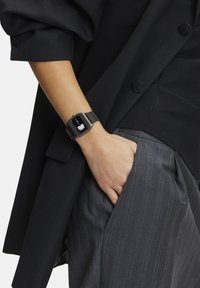 Rechthoekig smartwatch met een zwart matte display en een zilveren mesh band. Draag op een pols, gedeeltelijk bedekt door een donkere blazer met een gestructureerde afwerking.