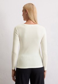 Haut à manches longues blanc à côtes, avec encolure ronde et silhouette ajustée. Texture douce avec des lignes verticales visibles et un ourlet droit.