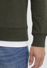 Dark green cotton sweater with a round neckline and long sleeves, featuring ribbed cuffs and a straight hem over a white shirt.