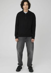 Pull noir à manches longues avec un col zippé, porté par-dessus une chemise de couleur claire, associé à un jean gris ample et des chaussures noires.