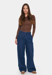 Ilga rankovėm ruda palaidinė, derinama su plačių kojų tamsiai mėlynomis džinsinėmis kelnėmis. Kelnės turi aukštą liemenį ir subtilią tekstūrą, kurias papildo rudi batai.