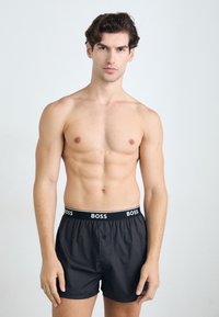 Boxer shorts noirs avec une ceinture logo BOSS, dotés d'une fermeture à bouton et d'un tissu lisse. Le modèle affiche une silhouette athlétique et ajustée.