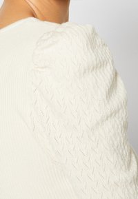 Haut blanc en maille à manches longues avec détails bouffants texturés aux épaules et motif chevron près de l’épaule.