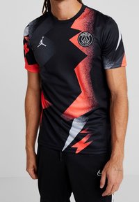 Man som bär en svart och röd Paris Saint-Germain fotbollströja med zigzag-mönster, står mot en neutral bakgrund.